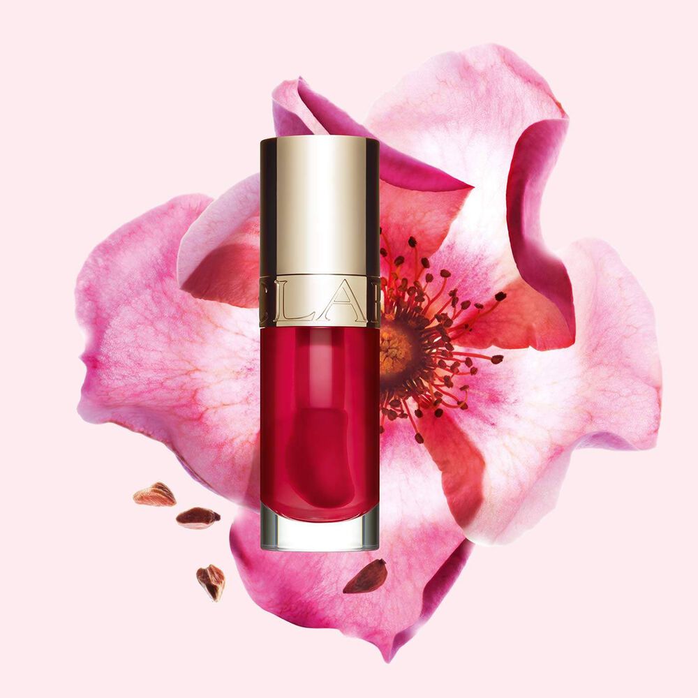 Packshot of Clarins Lip Comfort Oil Fushia, placed next to a sweetbriar rose on a light pink background.