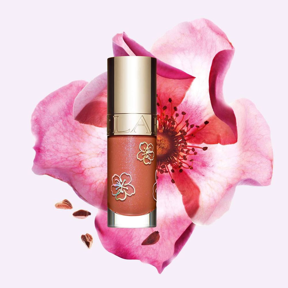 Packshot of Clarins Lip Comfort Oil in Rosy Sakura, a limited edition lip product in a pink shade, next to a flower.