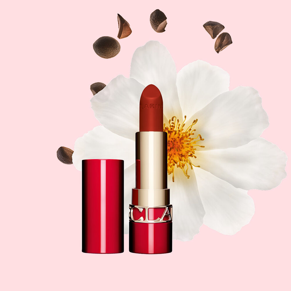 Packshot of Clarins matte Lipstick Joli Rouge in Shade 742 V, placed beside a camellia flower and shea seeds on a pink background.