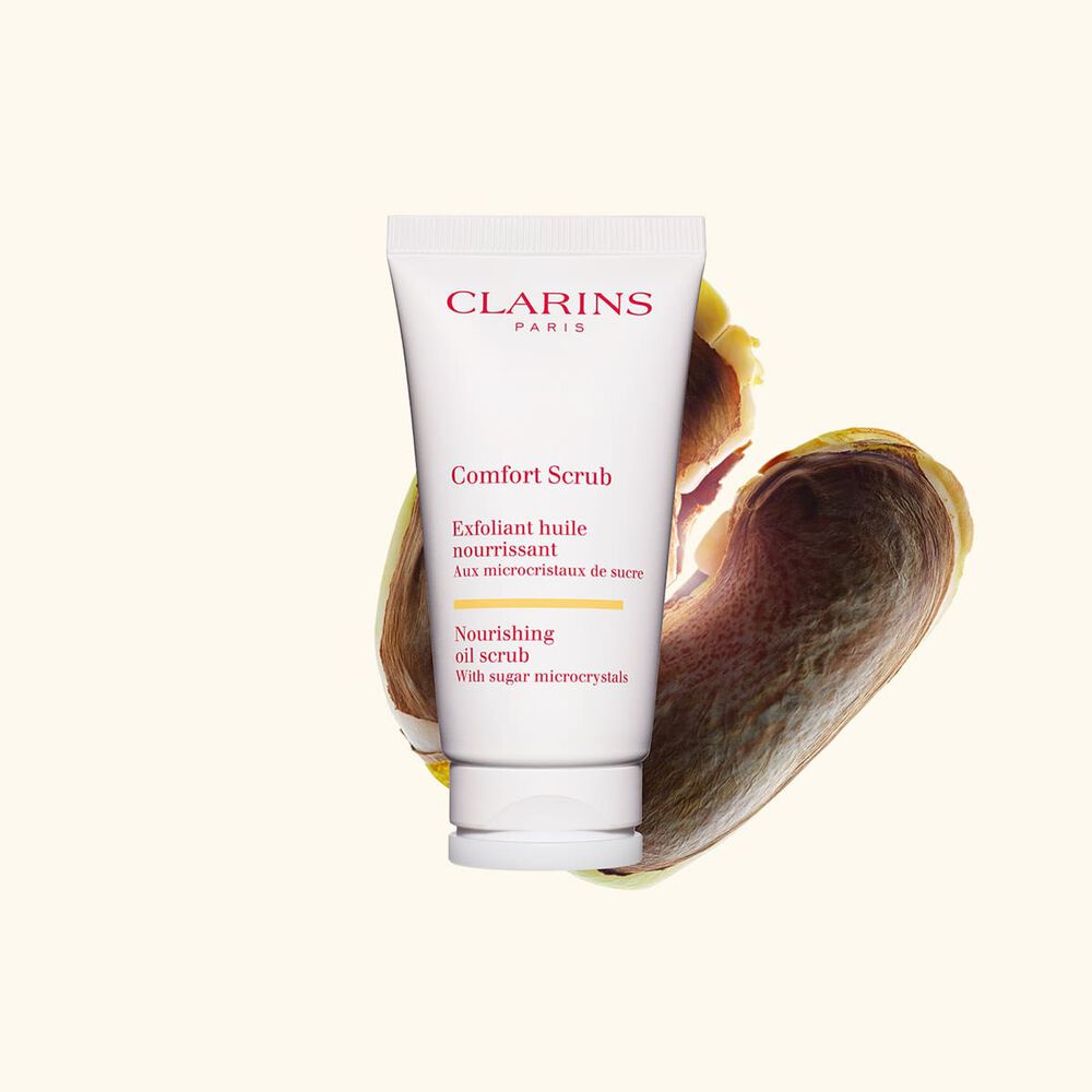 Packshot of Clarins face and lips exfoliant Comfort Scrub tube next to some wild mango seeds on a beige background