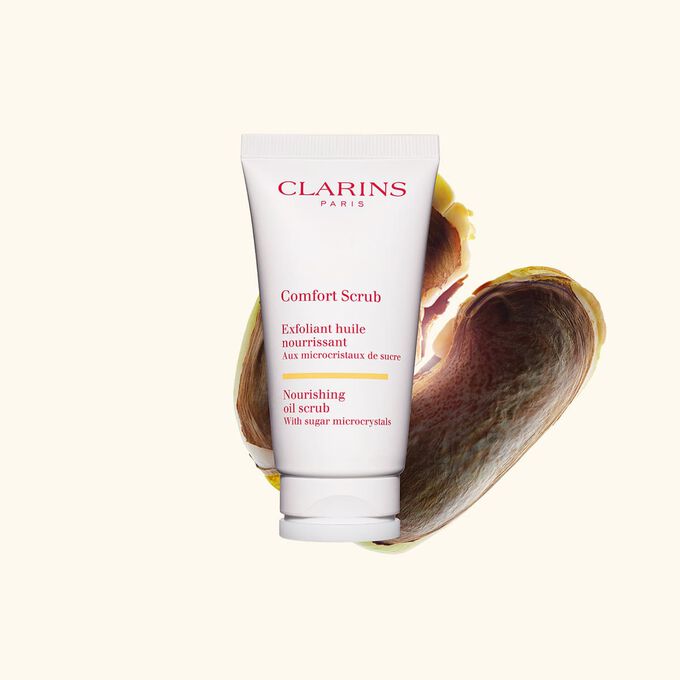 Packshot of Clarins face and lips exfoliant Comfort Scrub tube next to some wild mango seeds on a beige background