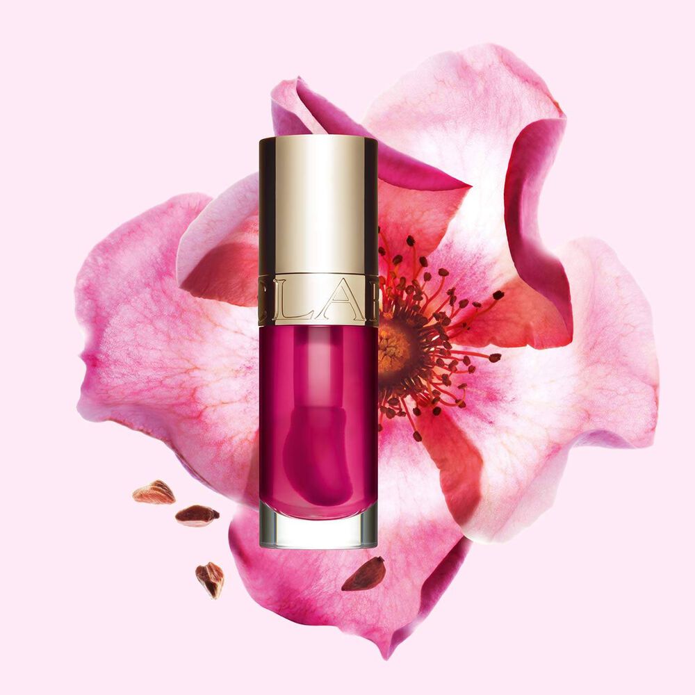 Packshot of Clarins Lip Comfort Oil in Raspberry, placed next to a sweetbriar rose on a light peach background.