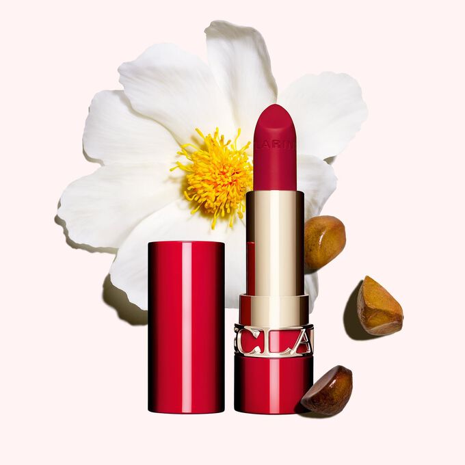 Packshot of Clarins matte Lipstick Joli Rouge in Shade 742 V, placed beside a camellia flower and shea seeds on a pink background.